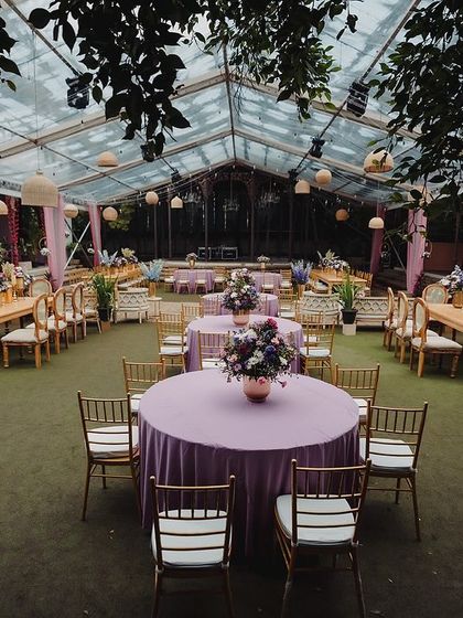 A beautiful pastel-themed Sangeet setup under a clear-top tent, with elegant table arrangements and hanging lanterns.