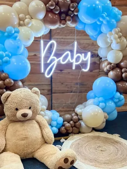 A circular balloon frame with a 'Baby' neon sign makes for a perfect photo booth, featuring a large, cuddly teddy bear.