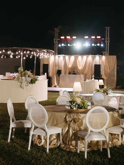 An elegant dinner setup for an evening soirée in Goa. The round tables are draped in gold fabric, with classic white chairs and soft lamp lighting creating a warm and inviting atmosphere.