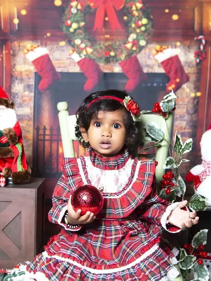 A classic Christmas portrait in front of a cozy fireplace. The toddler is dressed in a festive plaid dress, holding a shiny red ornament.