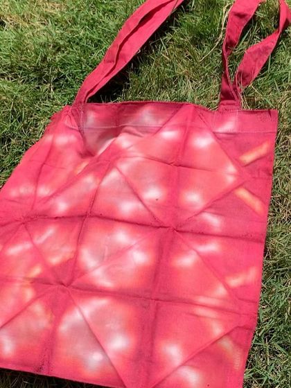 The same geometric shibori-dyed tote bag, laid out on the grass to dry. The red colour is vibrant and the pattern is crisp.