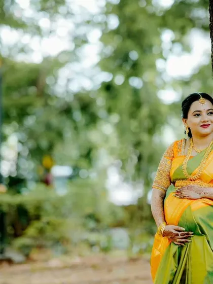 A thoughtful portrait of an expectant mother in a beautiful green and orange silk saree, leaning against a tree in a park. This image captures a moment of quiet reflection.