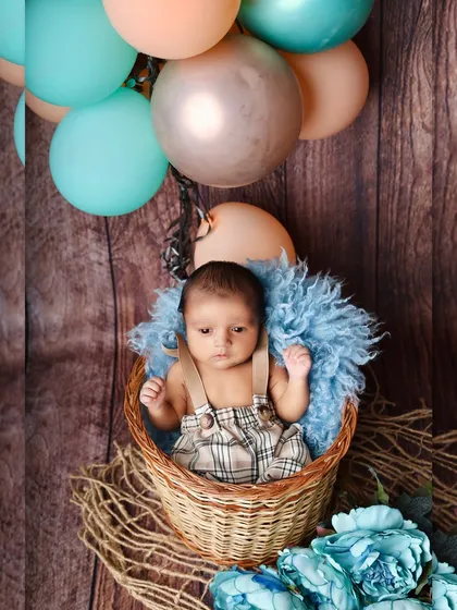 A cute baby boy in plaid overalls sits in a basket, surrounded by a hot-air balloon themed setup with blue and peach balloons.