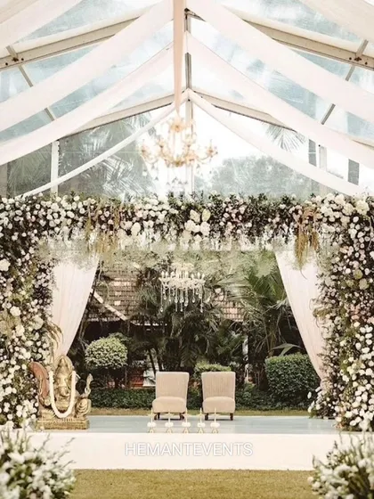 A beautiful wedding stage set inside a transparent tent, allowing for an outdoor feel with indoor comfort. The stage is heavily decorated with white flowers and features a Ganesha idol for blessings.