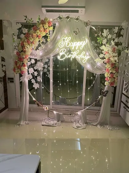Combining flowers and lights can create a truly dreamy effect. This beautiful birthday backdrop features a circular frame draped with sheer white fabric, elegant floral arrangements, and a glowing 'Happy Birthday' neon sign.