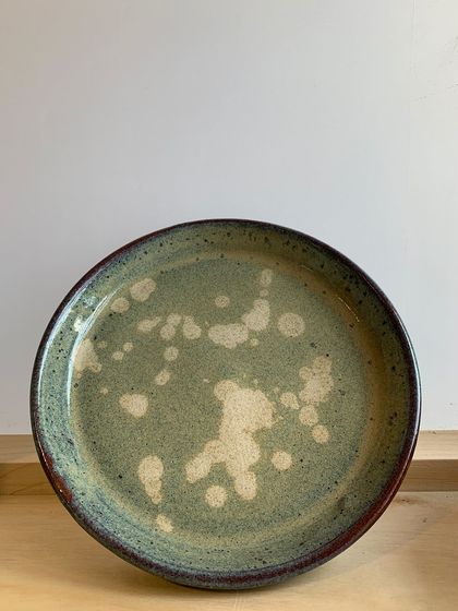 A handmade dinner plate with a beautiful, speckled green glaze. The abstract white splashes add a touch of modern artistry to this functional piece.