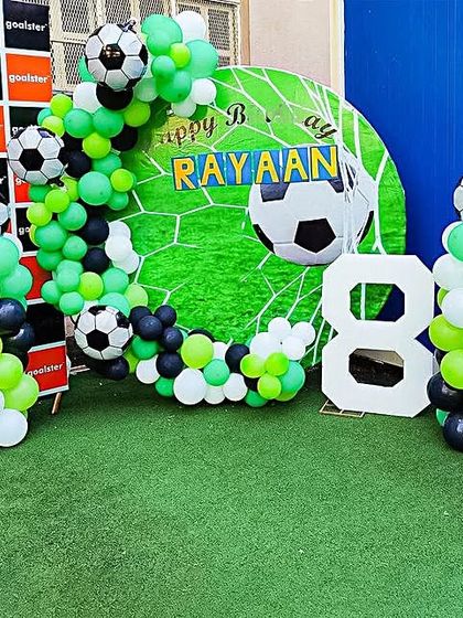 A football-themed party on an artificial grass surface. The decor includes a goal-themed backdrop, a green, black, and white balloon arch, and a large number '8'.