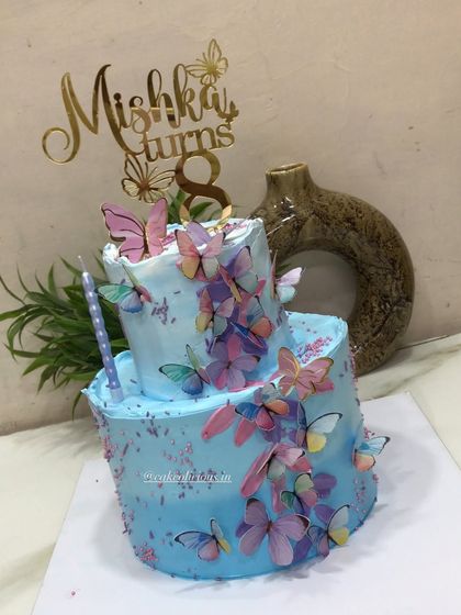 A pretty two-tier butterfly-themed cake for an 8th birthday. This 3kg chocolate mousse cake is covered in blue whipped cream and a cascade of colorful, edible butterflies, complete with a custom name topper.