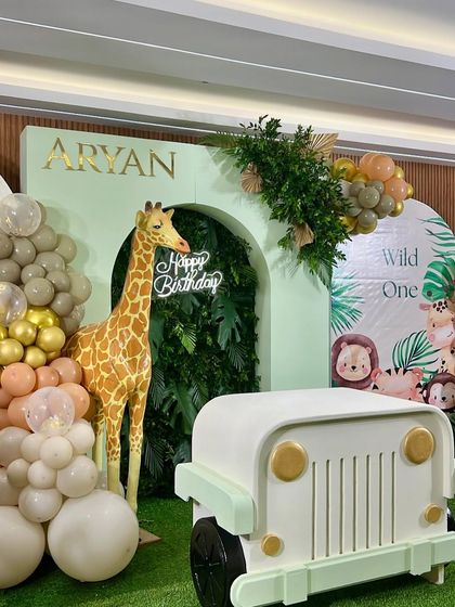 A closer look at the 'Wild One' jungle theme for Aryan's birthday. The setup includes a greenery wall with a neon 'Happy Birthday' sign, a life-sized giraffe, and a kid-sized jeep, perfect for photos.