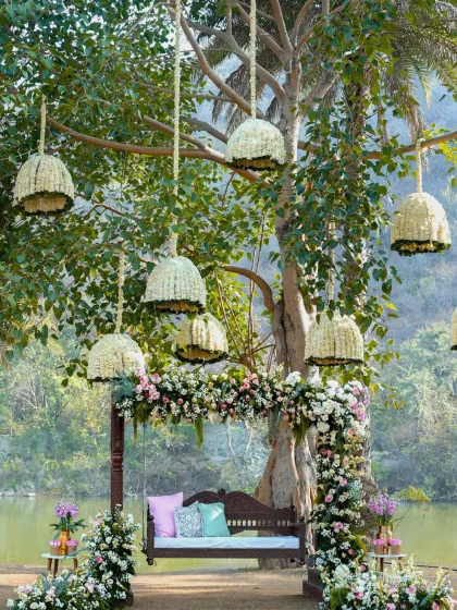 An enchanting Mehndi setup by a lake in Udaipur. A traditional wooden swing is decorated with pastel pink and white florals, while large floral chandeliers made of mogra hang from the tree above.