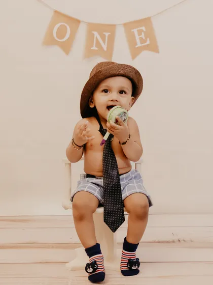 A cute studio shot of a one-year-old boy in a hat and tie, curiously inspecting a toy.