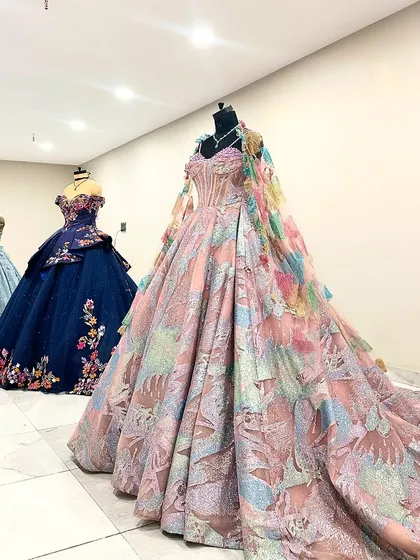 A view of our multi-colored ball gowns on display. The gown in the foreground features a stunning cape made of matching fabric, adding an extra layer of drama and elegance. In the background, you can see our navy floral and dusty blue gowns.