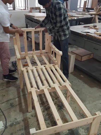 Diploma students collaborate on the assembly of a large lounge chair frame. Working together on complex projects teaches teamwork and problem-solving skills crucial in a professional environment.