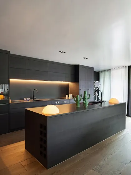 This modern pantry and breakout space was created for a corporate office, featuring sleek, handleless cabinetry and a large central island. Warm, under-cabinet lighting and natural wood floors balance the dark, minimalist aesthetic, providing a welcoming area for employees to connect.