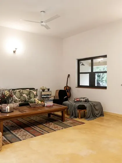 A relaxed, bohemian-style living room with a low wooden coffee table, a guitar in the corner, and plenty of green plants. The yellow-oxide flooring adds warmth and a rustic touch to the inviting space.