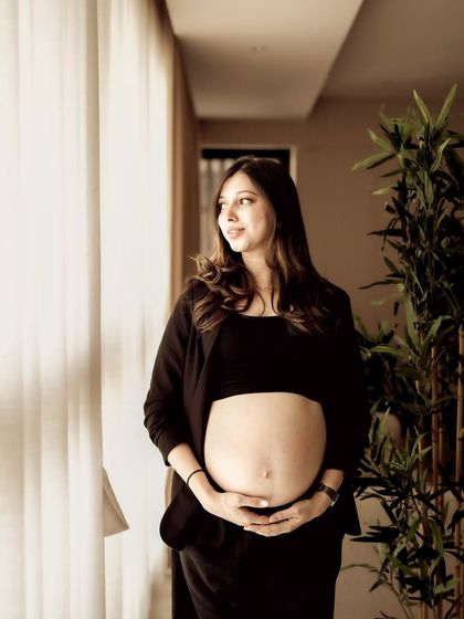 A beautiful window-lit indoor portrait. The mom-to-be looks out the window, her hands cradling her bare bump, in a moment of quiet thought.