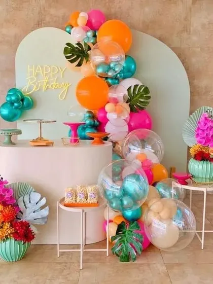A pop of tropical color for a vibrant birthday bash. This modern setup uses a unique color palette of orange, teal, and pink, with monstera leaf accents and clear balloons for a fun and stylish party.