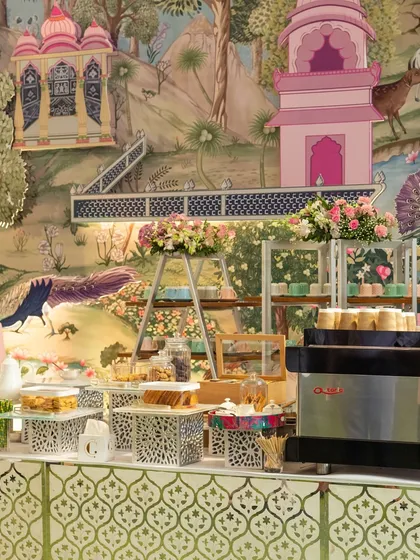 A beautiful high-tea setup with an espresso machine and an array of treats. The backdrop of whimsical, hand-painted wallpaper adds a unique and artistic touch to the coffee station.