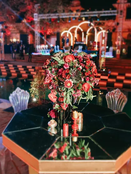 A centerpiece of rich red florals sits atop a mirrored, geometric table, reflecting the grand, neon-lit stage and adding a touch of moody romance to the seating area.