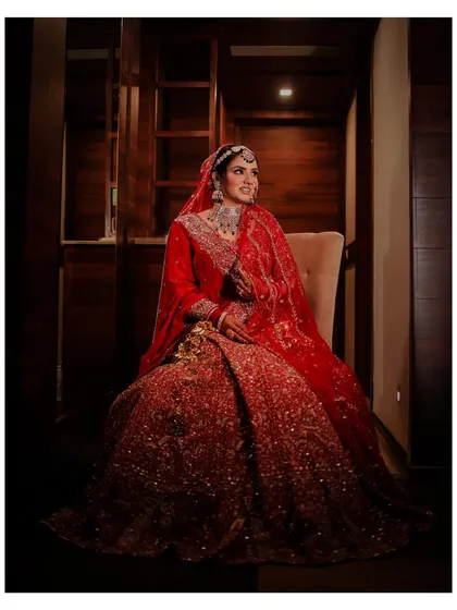A regal, seated portrait of the bride in her magnificent red lehenga. The dramatic lighting highlights the intricate embroidery of her outfit, creating a timeless and elegant image.