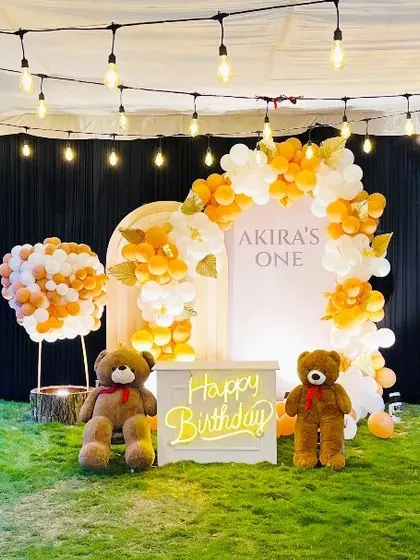 An adorable teddy bear themed birthday party set up outdoors on a lawn. The decor features two large teddy bears, a neon "Happy Birthday" sign, and an elegant balloon arch in white and orange.