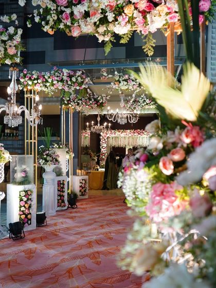 A stunning floral-draped walkway leading into the main event area. The path is lined with classic white pedestals and urns, creating a grand and romantic journey for guests.