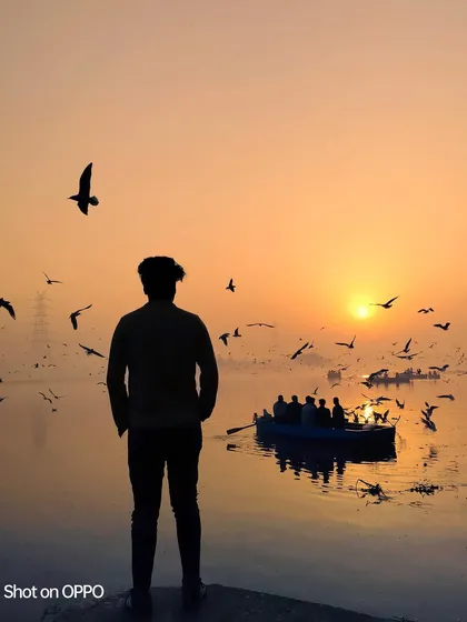 A silhouette of a person watching the sunrise over the Yamuna Ghat, as birds fly past and a boat full of people enjoys the view. It's a classic, beautiful morning scene.