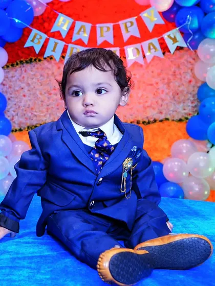 A toddler dressed in a sharp blue suit for his birthday photoshoot, set against a backdrop of blue and white balloons. We pay attention to the details of the decor and outfits.