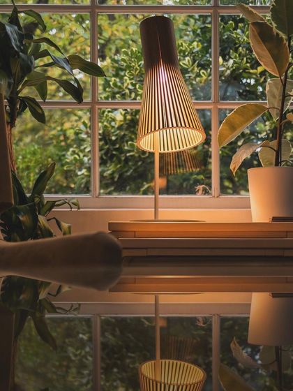 A Secto Design table lamp sits on a desk by a window, its slatted wooden shade casting a warm, gentle light that is perfect for reading or working.