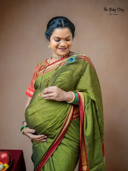 A close-up shot focusing on the mother-to-be's joyful expression and the details of her traditional attire. The peacock feather gently brushing her baby bump adds a symbolic and tender touch.