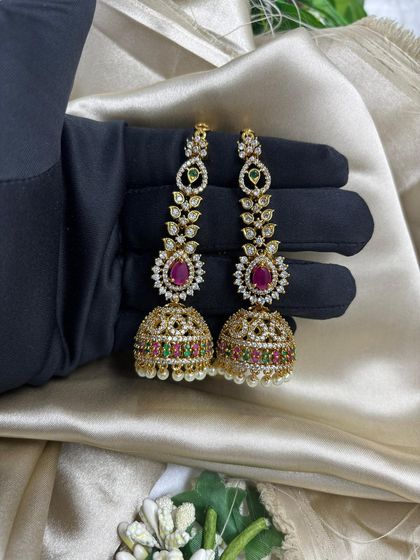A pair of traditional gold-plated jhumka earrings. These feature intricate work with AD stones and multi-colored accents, making them a timeless and essential item for any ethnic wear collection.