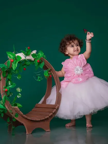 A whimsical shot of a toddler in a pink and white dress, interacting with a butterfly on a leafy prop. The dark green background makes the scene feel like a magical garden.