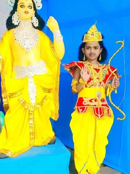 A young boy dressed as Lord Rama or Kartikeya, posing next to a deity during Durga Puja. The costume includes a decorated dhoti, chest plate, and crown, perfect for festival pandal hopping.