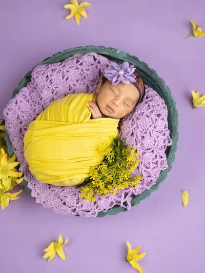 A vibrant and cheerful portrait. The baby is swaddled in bright yellow against a purple backdrop, surrounded by matching flowers.