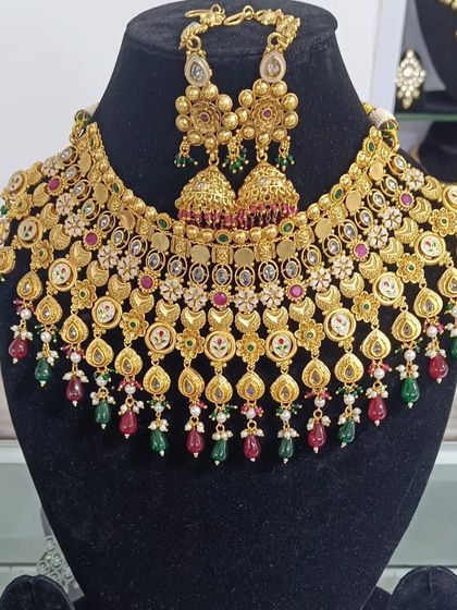 This heavy bridal choker set comes with matching jhumki earrings. The design features intricate gold-look work with red and green accents.