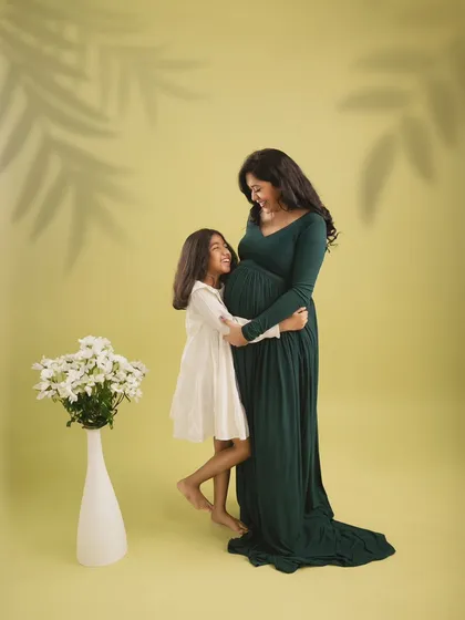 A simple and elegant studio maternity portrait. The mother-to-be and her daughter share a loving gaze, framed by soft lighting and the subtle shadow of a palm leaf.