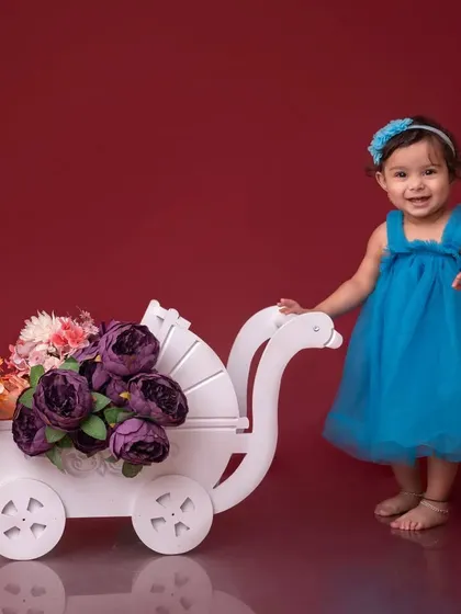 A sweet little girl in a blue dress pushes a white pram filled with flowers. This setup against a red background is perfect for a colorful and charming portrait.