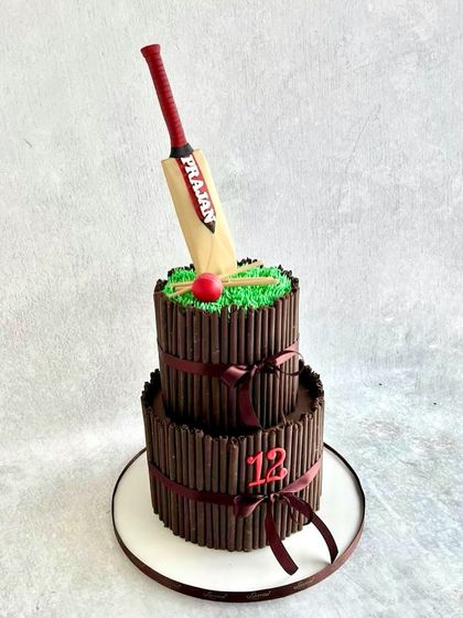 A cricket-themed cake for Prajan's 12th birthday. This two-tiered cake is covered in chocolate sticks to look like a wicket, with a fondant cricket bat and ball on top.