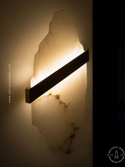 This artistic wall sconce features a raw, organically shaped slab of translucent marble. A simple diagonal metal bar houses the light, which glows through the stone to reveal its natural veining and character.