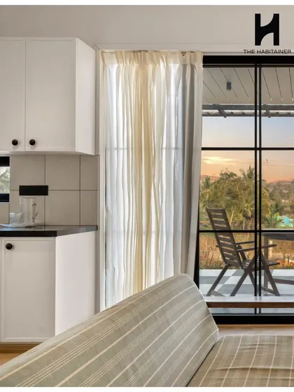 A view from the living room of the French-themed villa, looking out onto the deck. The clean white modular kitchen and light herringbone flooring contribute to the bright and airy feel of the space.