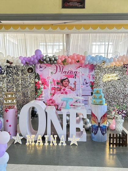 A grand butterfly theme in a hall, featuring a silver shimmer wall and personalized backdrops with the birthday girl's photos. The large 'ONE' marquee letters and pastel balloon arch complete this elaborate first birthday stage.
