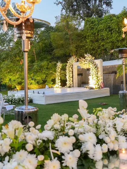 A view of the stunning outdoor setup for the anniversary, with a white floral stage, elegant seating, and chandeliers creating a magical garden party atmosphere.