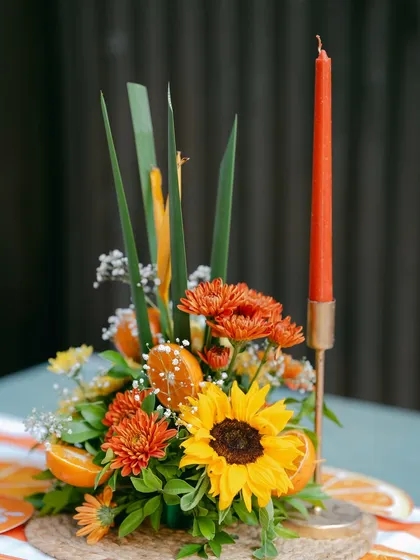 A vibrant table centerpiece with sunflowers, orange chrysanthemums, and slices of fresh tangerine, complete with a taper candle.