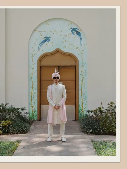 A full-length portrait of the groom looking sharp and stylish in his wedding attire, standing before a beautifully painted archway.