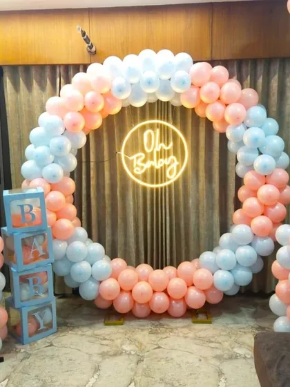 A classic 'gender reveal' style baby shower decoration for the home. It features a full balloon ring in pastel pink and blue, an 'Oh Baby' neon sign, and 'BABY' blocks on either side.