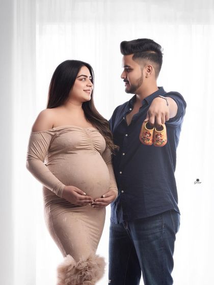 A sweet and classic couple pose, with the dad-to-be holding up a pair of baby shoes. The neutral-toned outfits and bright background create a warm and happy feeling.
