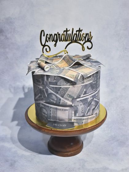 A cake that looks like a stack of money. This unique design is wrapped in edible Indian currency notes and topped with a "Congratulations" sign, perfect for celebrating a big success.