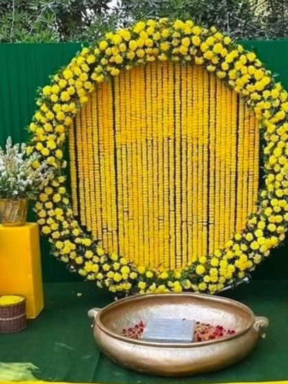 A classic Haldi backdrop for outdoor functions. This setup features a large circular frame filled with strings of yellow marigolds, a green panel background, and a traditional brass urli in the center for the ceremony.