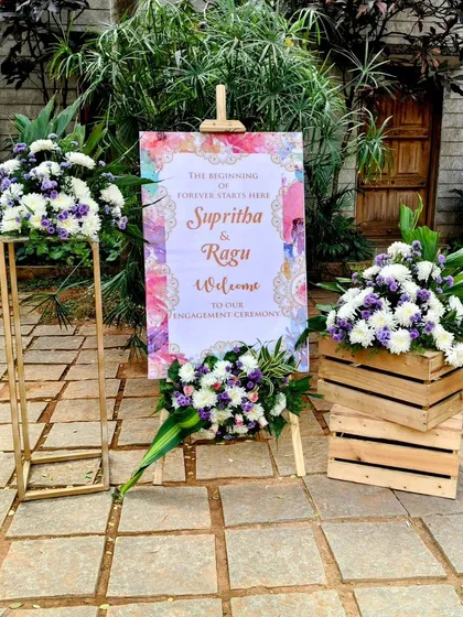 The small details matter. This custom welcome sign for an engagement ceremony, framed by fresh floral arrangements on rustic wooden crates and a sleek gold stand, sets a personal and inviting tone right from the entrance.