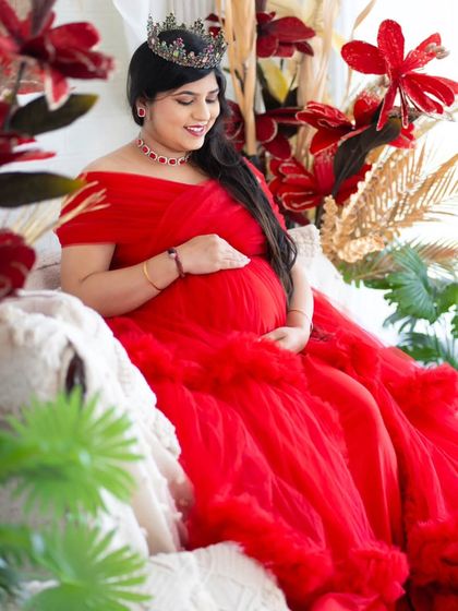 The queen of the hour. This mom-to-be looks radiant in a bold red gown, seated comfortably in our boho studio setup surrounded by vibrant floral arrangements.